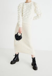 Pull en maille blanc avec manches bouffantes et motif torsadé, associé à une jupe midi blanche en fausse fourrure. Sac à main noir en forme de cœur et bottines épaisses noires.