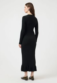 Μαύρο ριμπ πλεκτό φόρεμα με μακριά μανίκια, εφαρμοστή σιλουέτα και βολάν στο τελείωμα. Το μοντέλο φοράει σκούρα επίπεδα παπούτσια. Ομαλή υφή.