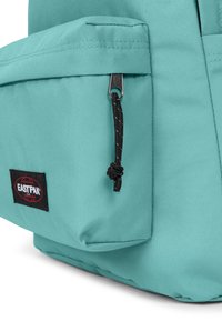 Turkos ryggsäck i slitstarkt tyg, med en liten sidoficka, en svart dragkedja med flätad dragsko och en etikett med logotyp.