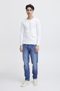 Fehér, hosszú ujjú ing, kék farmer, világos textúrák, laza szabás, krémszínű sportcipő, egyszínű fehér háttér előtt állva.