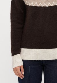 Nærbilde av en person som har på seg en mørkebrun genser med kremfargede mansjetter og søm, blå jeans og en mønstret beige yoke.