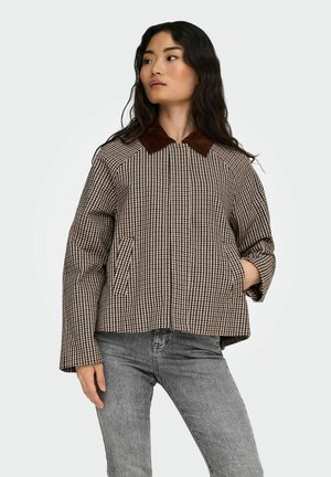 Femme portant une veste à carreaux marron et beige avec un col marron foncé et un jean gris, debout et regardant vers sa gauche.