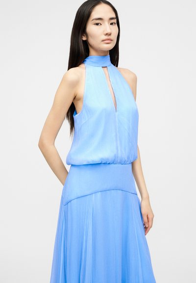 Νεαρή γυναίκα με μακριά σκούρα μαλλιά, που φοράει αμάνικο φωτεινό γαλάζιο φόρεμα με λαιμόκοψη halter και άνετη φούστα.