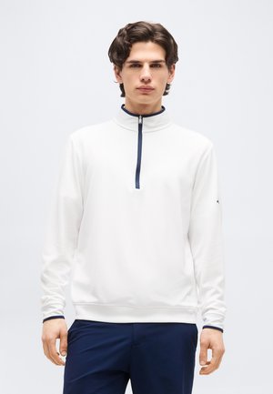 Ung mann som har på seg hvit genser med halv glidelås og marineblå kant, og marineblå bukser, står foran en ensfarget lys grå bakgrunn.