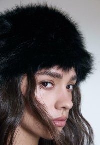 Young woman with long dark hair wearing a black furry hat, looking sideways against a plain light background.