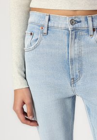 Jeans en denim bleu clair avec une taille haute, surpiqûres contrastées, poches avant et une texture légèrement délavée. Le bouton et les rivets sont de teinte cuivrée.