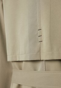 Gros plan sur le tissu beige d’un trench-coat montrant des coutures détaillées et des couches superposées avec une ceinture à la taille.