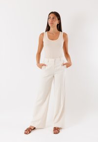 Top beige abbinato a pantaloni cremosi a gamba larga con vita alta, dotati di tasche laterali e un tessuto liscio e testurizzato. Sandali marroni completano il look.