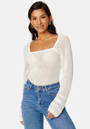Weißer Strickpullover mit langen Ärmeln, der über einen quadratischen Ausschnitt und ein netzartig wirkendes Muster verfügt. Kombiniert mit hochgeschnittenen blauen Jeans.