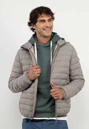 Hombre sonriendo mientras lleva una sudadera con capucha verde bajo una chaqueta acolchada gris claro, de pie frente a un fondo blanco liso.