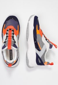 Pár vícebarevných sportovních tenisek s oranžovými, námořnickými, bílými a šedými akcenty, zobrazující vrchní a boční pohled na bílém pozadí.