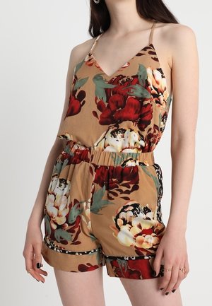 Nainen pukeutuneena beigeen kukkakuosiseen hihattomaan haalaripukuun, jossa on punaisia, valkoisia ja vihreitä kukkia sekä joustava vyötärö.