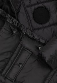 Black puffer jacket with quilted texture, featuring a round logo patch, zip closure, and snap buttons on the front pocket.