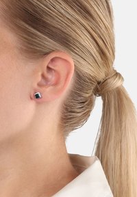 Närbild av en person med blont hår som är uppsatt och bär en liten svart fyrkantig sticköre i vänster öra.