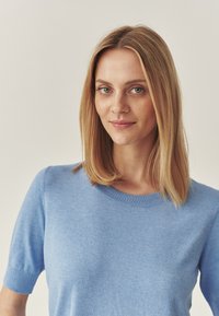 Pulover scurt, de culoare albastru deschis, cu mânecă scurtă și decolteu rotund, realizat dintr-un material tricotat moale. Textură netedă, siluetă ajustată.