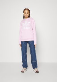 Rosa hettegenser med hvit adidas-logo, kombinert med blå denimbukser og beige sneakers med brune aksenter.