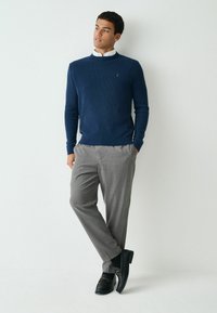Marineblauer Strickpullover, graue, taillierte Hose, schwarze Slipper mit Schnallen-Detail. Modell präsentiert ein lässiges, aber zugleich elegantes Outfit.