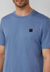 Lichtblauw katoenen T-shirt met een ronde hals, korte mouwen en een klein zwart vierkant logo-patch op de linkerborst.