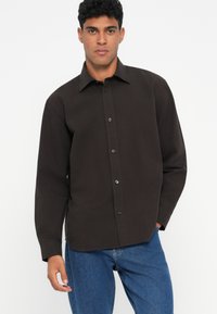 Mørkebrun skjorte med knappestolpe og krage, lange ermer og en glatt tekstur, kombinert med denimjeans.