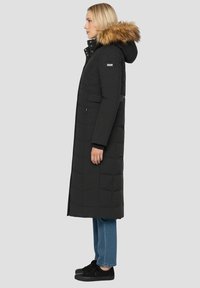 Długa czarna pikowana kurtka puchowa z kapturem obszytym futrem, z pikowanym wzorem, bocznymi kieszeniami i zapięciami na guziki, noszona na niebieskich dżinsach i czarnych butach.