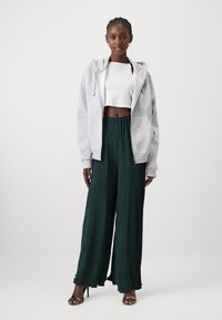 Szürke, zipzáras kapucnis pulóver, fehér cropped top és zöld, redőzött, bő szárú nadrág. A modellen fekete magassarkú szandál van. Simás textúrák és laza szabás.