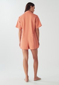 Persikofärgad kort overall med krage, korta ärmar och två bakfickor. Mjukt tyg och avslappnad passform.