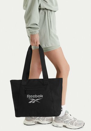 Persona con shorts y sudadera Reebok de color verde claro, sosteniendo una bolsa tote negra de Reebok, con zapatillas Reebok blancas y grises visibles.