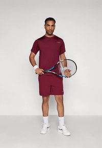 Homem em traje atlético bordo segurando uma raquete de ténis, vestindo pulsozeiras brancas, meias e ténis brancos, de pé contra um fundo liso.