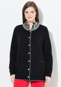Femme portant un cardigan noir à boutons avec une bordure grise, une chemise à col fleuri, un pantalon rouge et des boucles d'oreilles en or, se tenant devant un fond blanc.