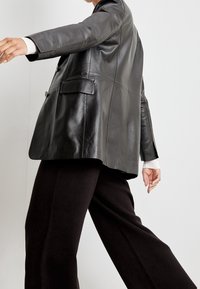 Czarna skórzana marynarka o luźnym kroju, z pojedynczym zapięciem na guzik i dwoma bocznymi kieszeniami. Gładka tekstura, czyste linie i subtelny połysk.