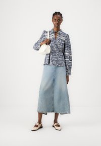 Blå blomstret bluse med lange ermer og krage, paret med en lys blå denimmaxikjole. Hvitt håndbagasje og beige flate sko.