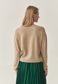 Beige gebreide trui met lange mouwen, ronde halslijn en ribbelzoom, gecombineerd met een groene geplooide rok. Gladde textuur zichtbaar.