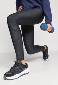 Μαύρο κολάν με διακριτικό σχέδιο φύλλων, συνδυασμένο με μαύρα και λευκά αθλητικά παπούτσια. Κρατάει έναν μπλε αλτήρα σε τεταμένη στάση.