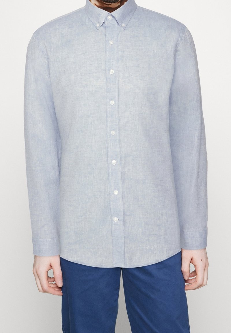 Lichtblauw linnen overhemd met lange mouwen en knopen. Heeft een spitse kraag en witte knopen, met een licht gestructureerde stof.