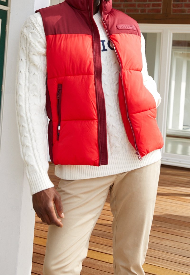 Persona vistiendo un chaleco de plumas rojo y burdeos sobre un suéter blanco de punto trenzado y pantalones beige, de pie en una terraza de madera cerca de una puerta.