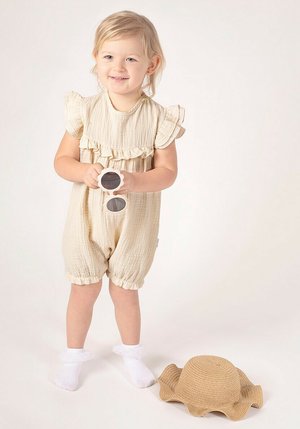 Beżowy, teksturowany bawełniany kombinezon z falbanianymi rękawkami i elastycznymi mankietami. Obok białe skarpetki i słomkowy kapelusz przeciwsłoneczny z falbanką na brzegu. Dziecko trzymające okulary przeciwsłoneczne.