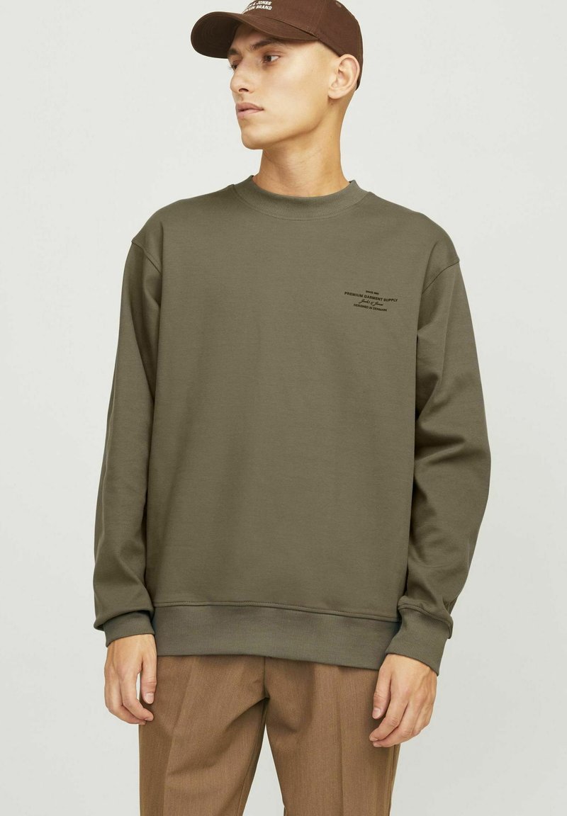 Olíva zöld pulóver ribbelt mandzsettákkal és aljjal, a mellkason kis fekete nyomtatással. Barna nadrággal és hozzá illő sapkával párosítva.