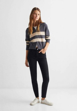 Gebreide cardigan in marineblauw met beige strepen, cropped ontwerp, geribbelde randen. Draagt met zwarte skinny jeans en witte sneakers.