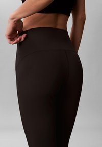 Γυναίκα που φοράει μαύρο κολάν ψηλόμεσο, τραβώντας ελαφρώς τη μέση του κολάν στο αριστερό της ισχίο, μπροστά σε απλό γκρι φόντο.