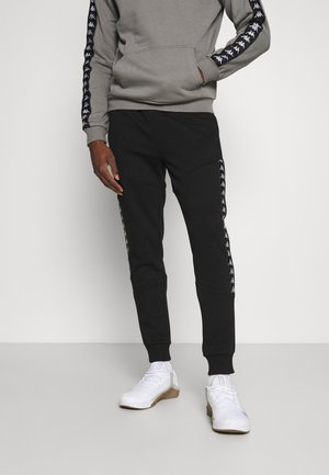 Szara bluza z kapturem i kieszenią typu kangur, w połączeniu z czarnymi spodnie dresowymi z bocznymi paskami oraz białymi butami sportowymi.