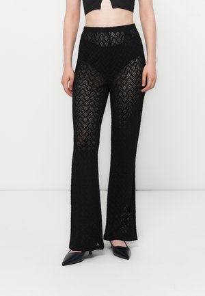 Frau trägt hoch taillierte schwarze Spitzenhose mit Zickzackmuster, kombiniert mit schwarzen spitzen Pumps und einem kurzen schwarzen Top.