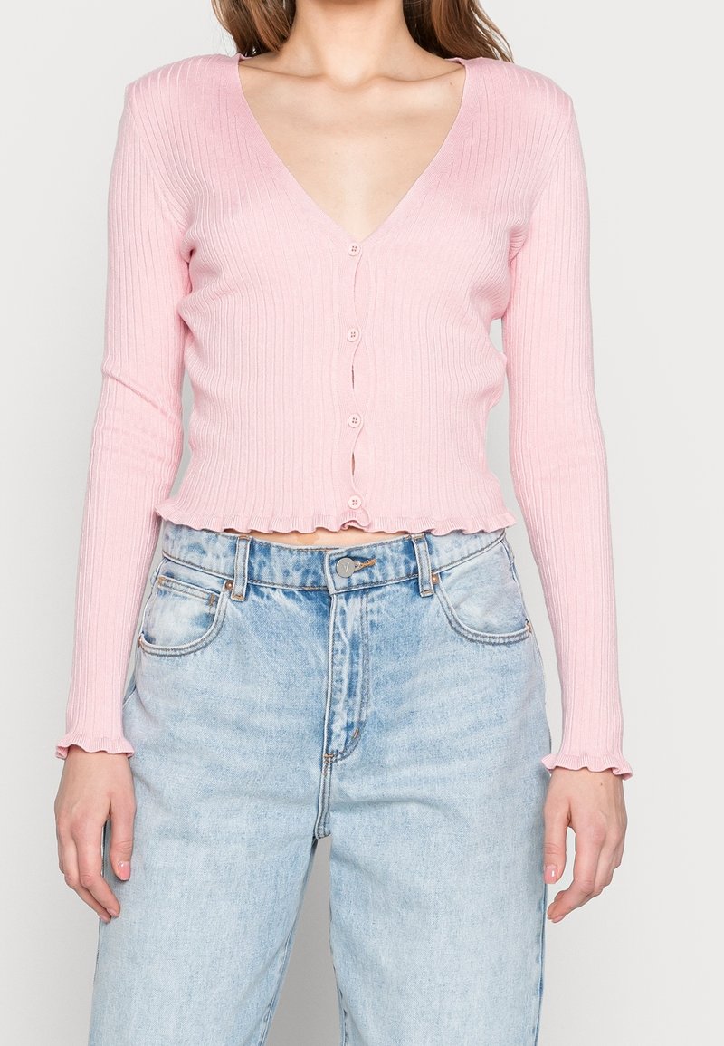 Helles rosa geripptes Cardigan mit langen Ärmeln, V-Ausschnitt und Knopfverschluss. Merkmale sind sanft gerüschter Saum und Manschetten. Getragen mit hochrising Jeans.