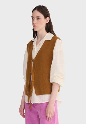 Kobieta ubrana w musztardową dzianinową kamizelkę zawiązaną z przodu, na białej bluzce z długim rękawem i różowych spodniach, stojąca na jednolitym tle.