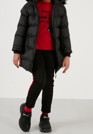 Enfant portant une doudoune noire par-dessus une chemise rouge, un pantalon noir avec une bande rouge, des chaussettes rouges et des baskets noires, debout sur un sol gris.
