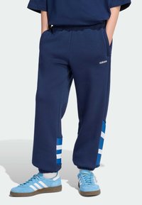 Tmavomodré teplákové nohavice s modro-bielymi pruhovanými akcentmi na manžetách. Majú elastický pás a bočné vrecká. Zobrazené je logo Adidas.