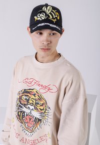 Beige sweatshirt med tiger grafik och "Ed Hardy" text, tillsammans med en svart keps med en tecknad bi-design och mesh bak.