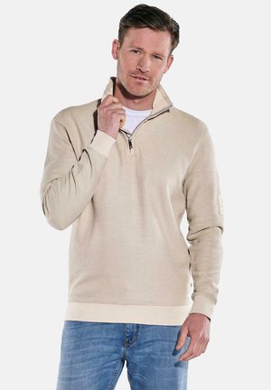 Beige Zip-Hoodie aus strukturiertem Stoff, mit hohem Kragen, gerippten Ärmeln und kleinem Logo auf der Brust. Kombiniert mit hellblauen Jeans.