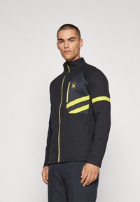 Veste zippée noire avec un col montant, présentant des accents jaunes, un tissu texturé et une poche poitrine gauche avec des rayures.