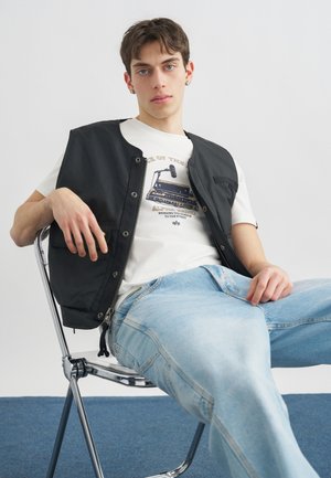 Jonge man zittend op een metalen stoel, gekleed in een zwart vest, een wit T-shirt met print en lichtblauwe spijkerbroek, tegen een effen achtergrond.