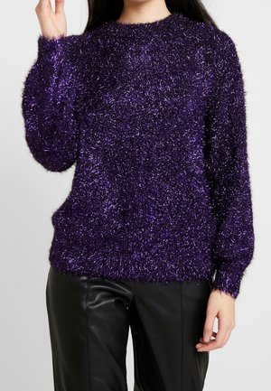 Femme portant un pull violet duveteux texturé brillant et un pantalon en cuir noir, une main levée près de sa tête.